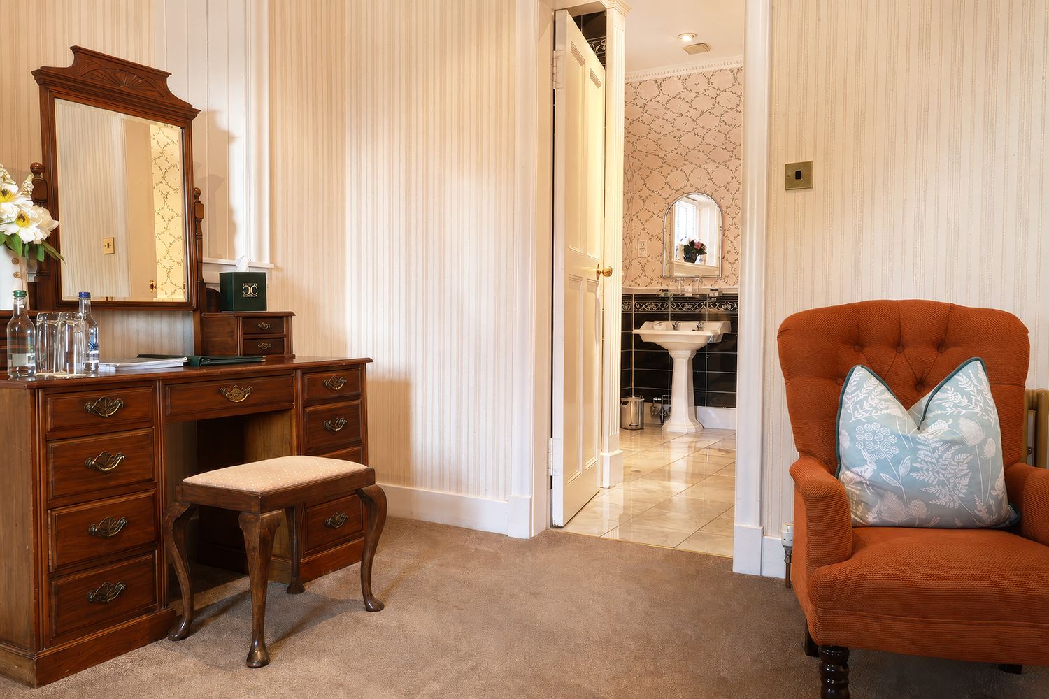 A room with a wooden dresser, a chair with a floral cushion, and a bathroom visible through an open door.