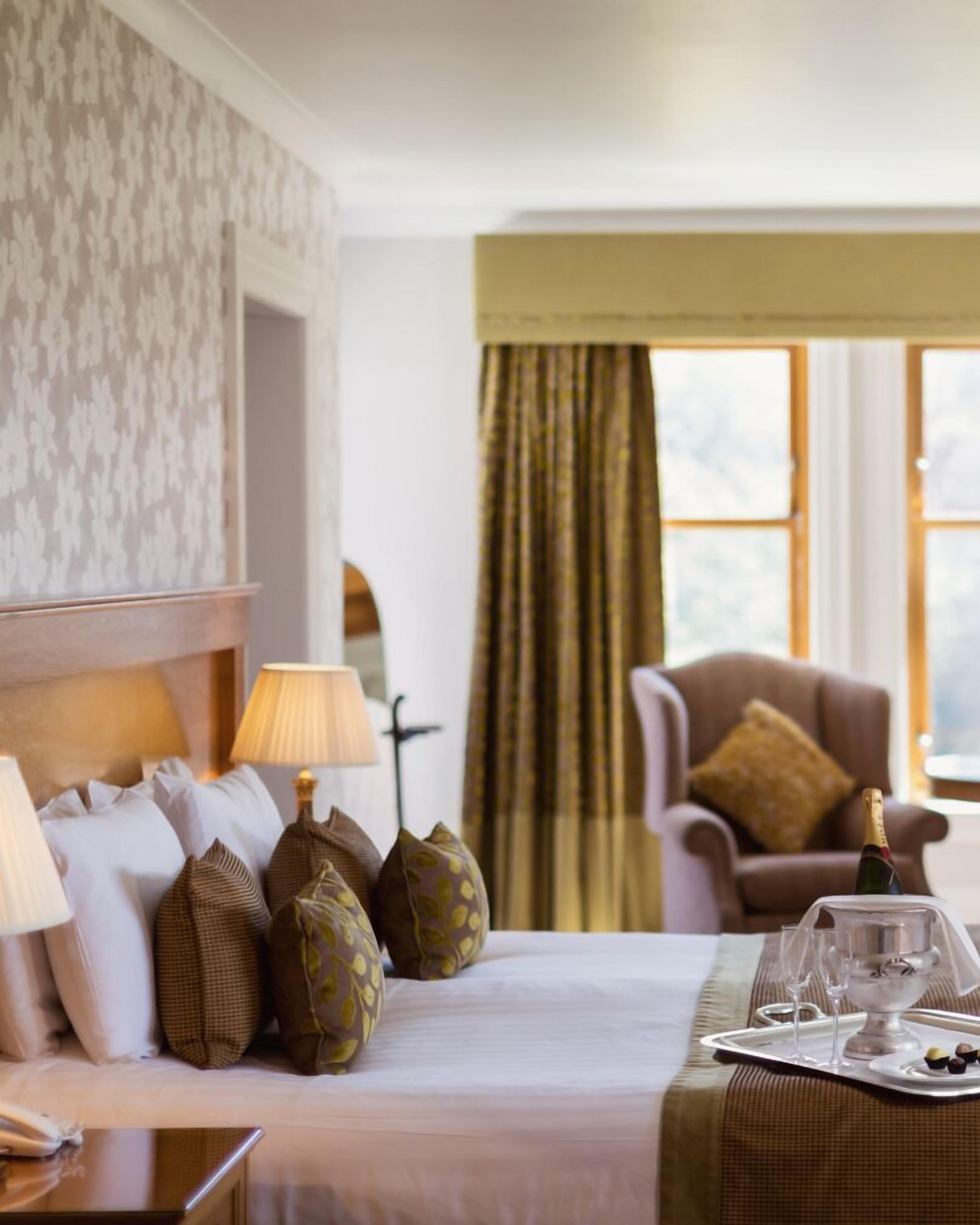 A hotel room with a neatly made bed, a chair, a table with a tray of glasses and snacks, and a view of the outdoors through the window.
