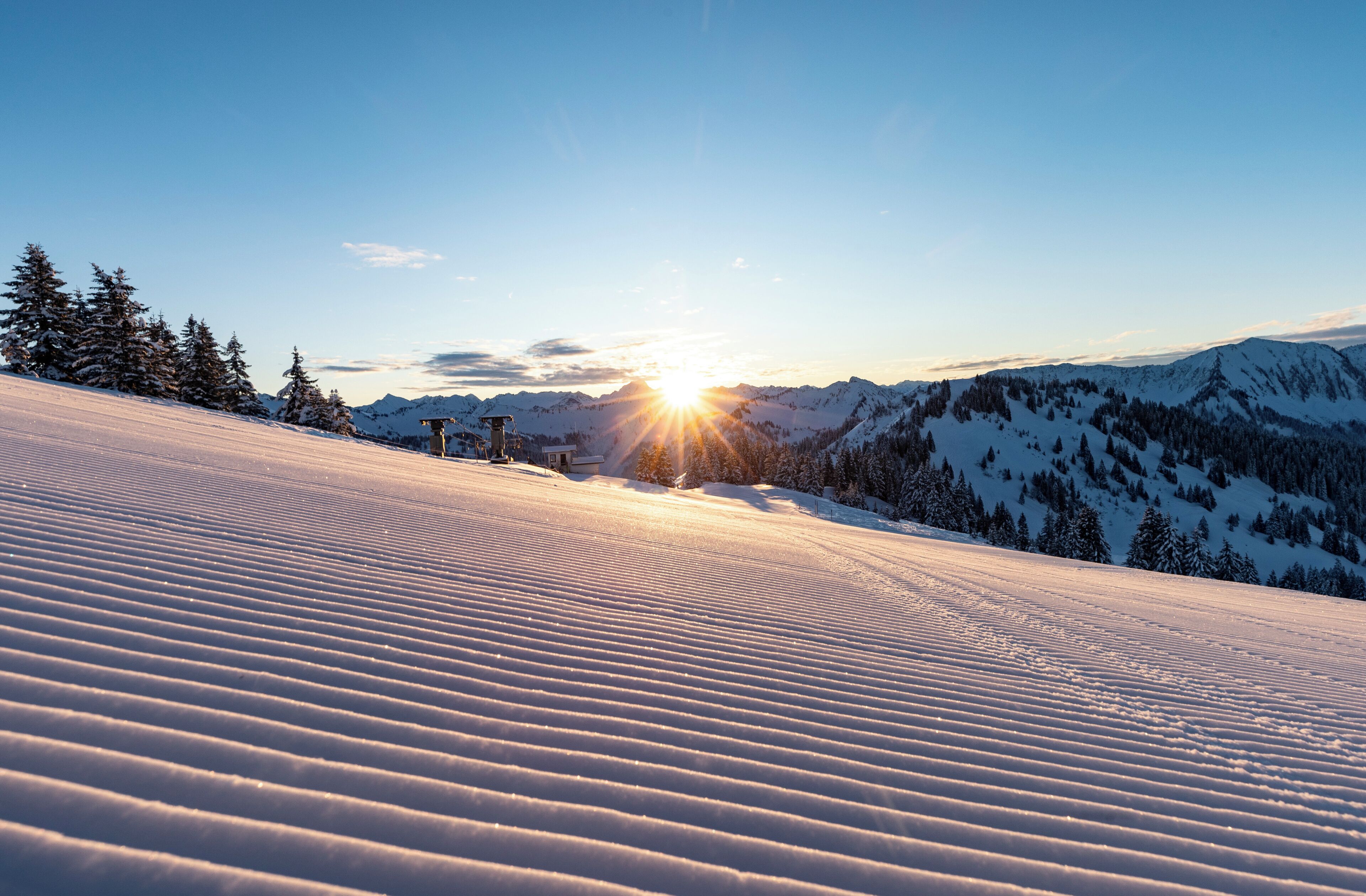 A snow-covered mountain slope with a clear sky and the sun setting or rising in the background.