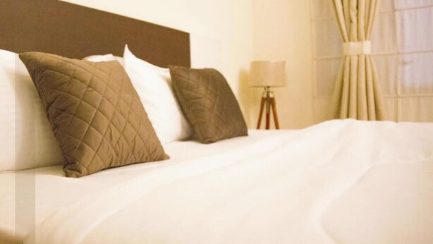 A hotel room with a neatly made bed, a lamp on a side table, and a window with curtains.