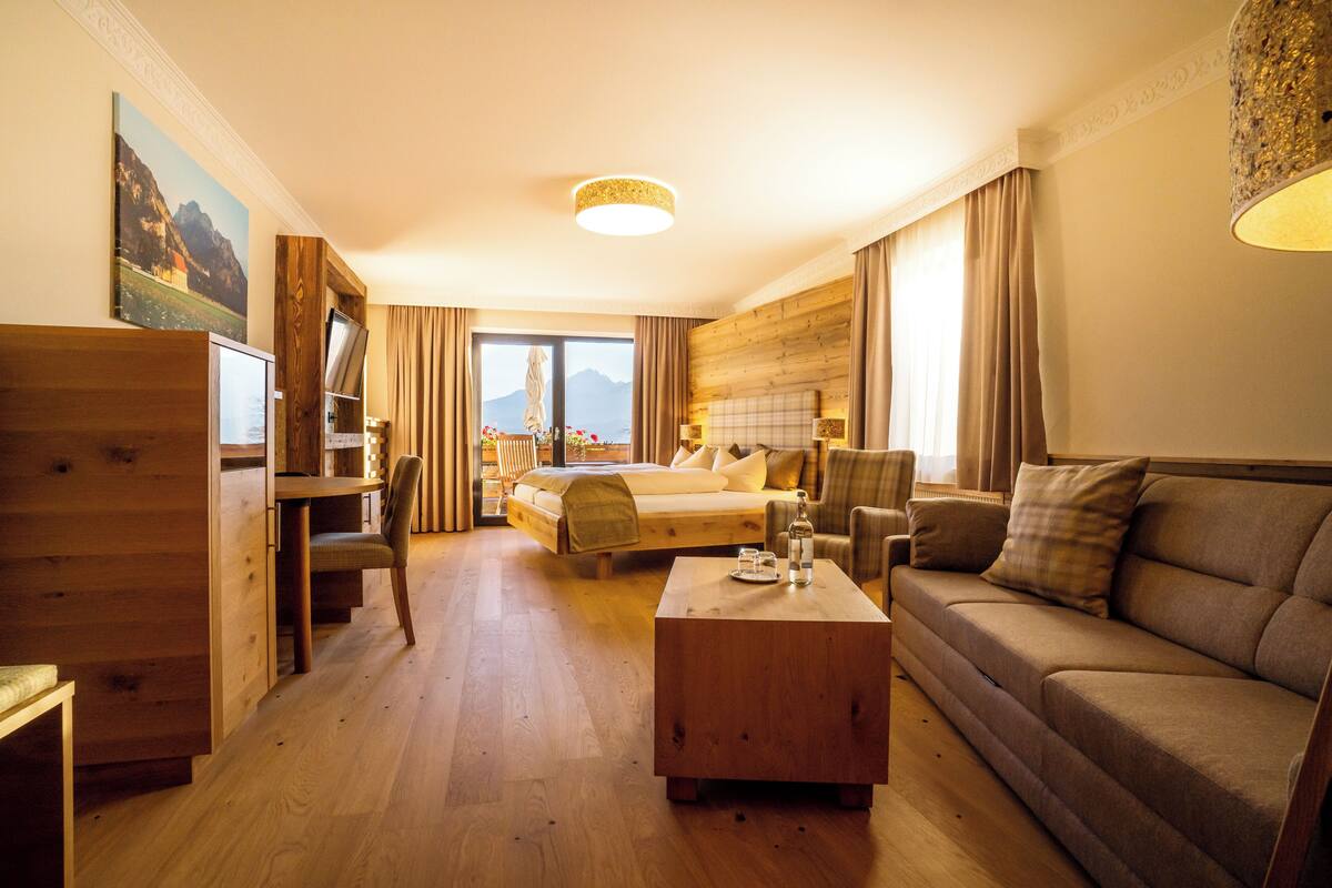 Een hotelkamer met een houten vloer, een bed met uitzicht op de bergen, een bank, een salontafel en een bureau met een stoel.