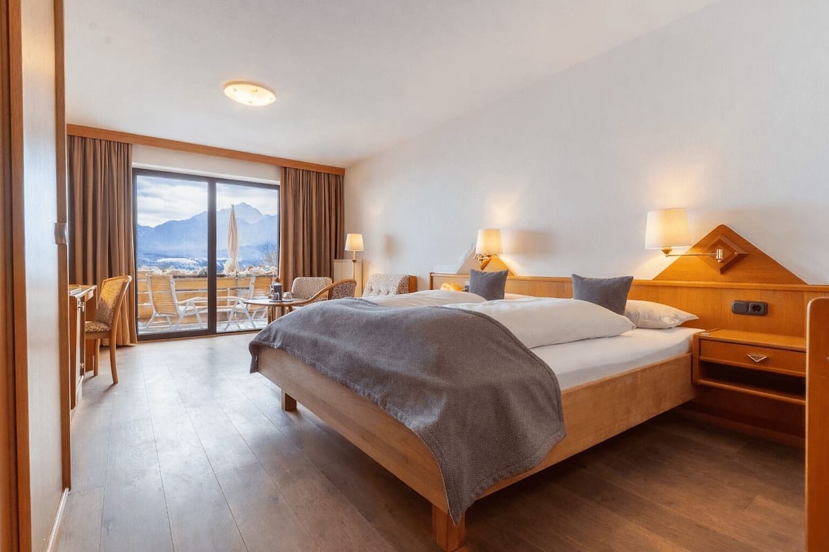 Hotelkamer met een groot bed, twee nachtkastjes, een stoel, een balkon met uitzicht op bergen en een houten vloer.