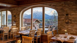 A restaurant with wooden furniture and large arched windows offering a mountain view. - Biohotel Eggensberger (Fuessen)