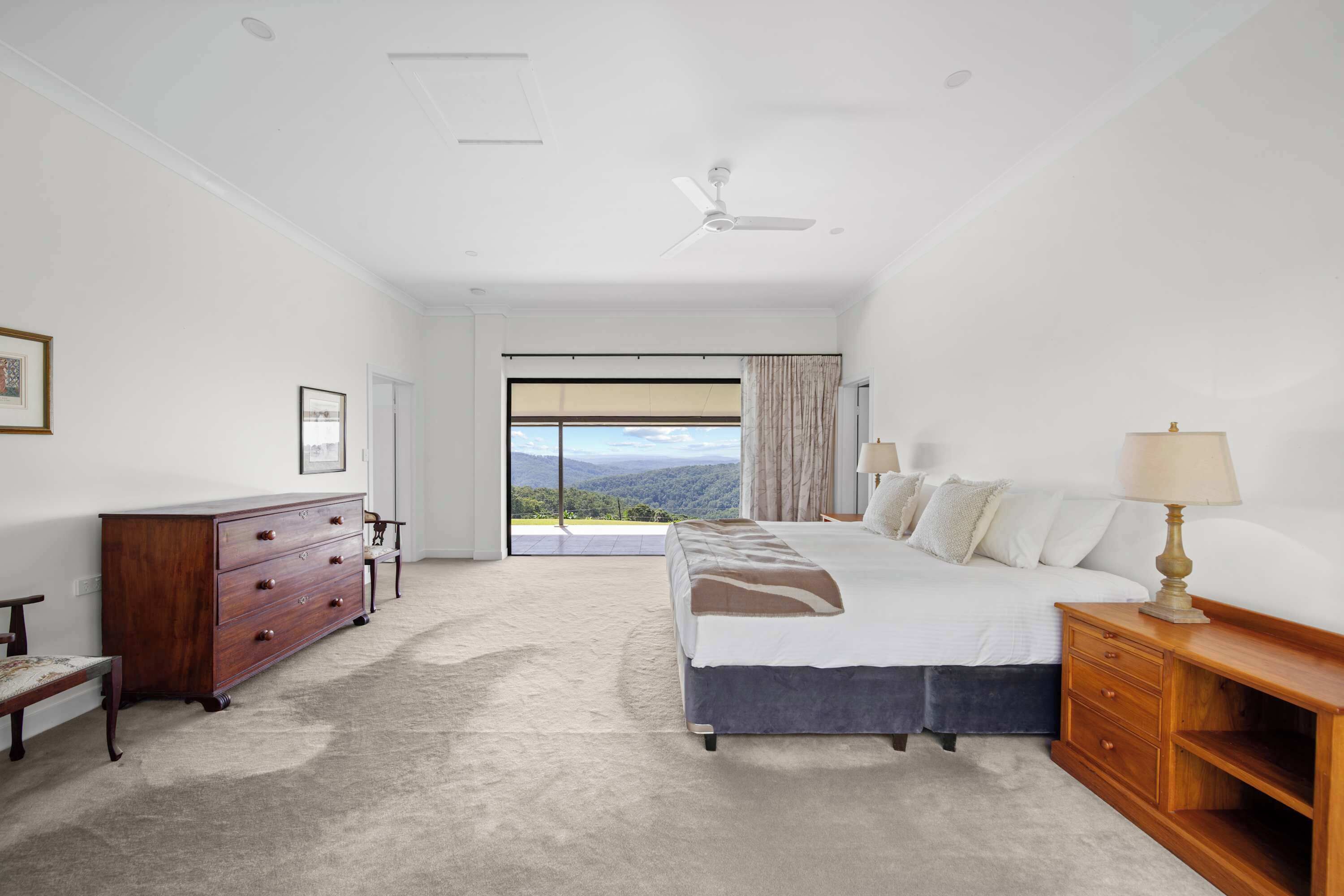A bedroom with a large bed, a dresser, a nightstand, a ceiling fan, and a view through a large window.