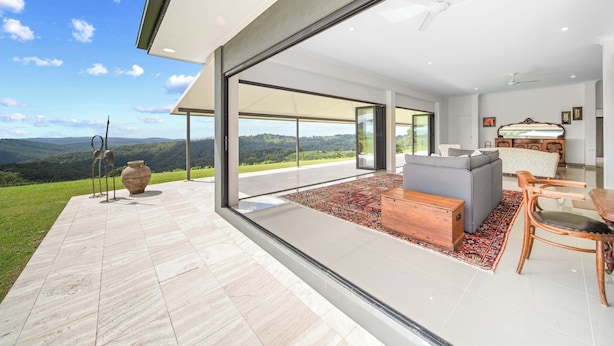 A modern interior with a large glass door leading to a balcony, a grey sofa, a wooden chest, and a dining table with chairs. - Panorama Farm - 3BD Wilderness Retreat (Hinterland Region)