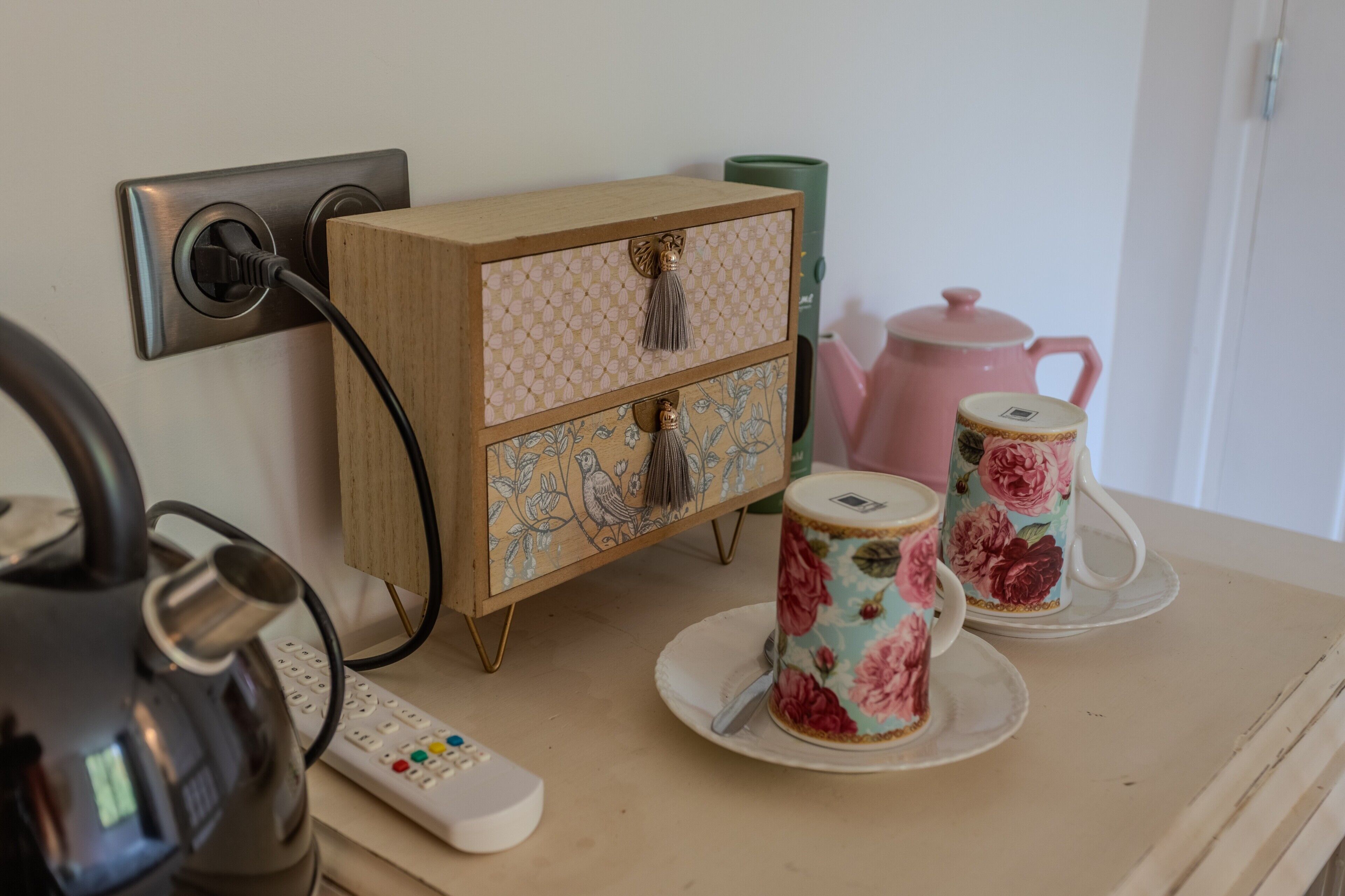 Een kitchenette met een waterkoker, een afstandsbediening, een houten kastje met decoratieve panelen en bloemendecor cups op een dienblad.