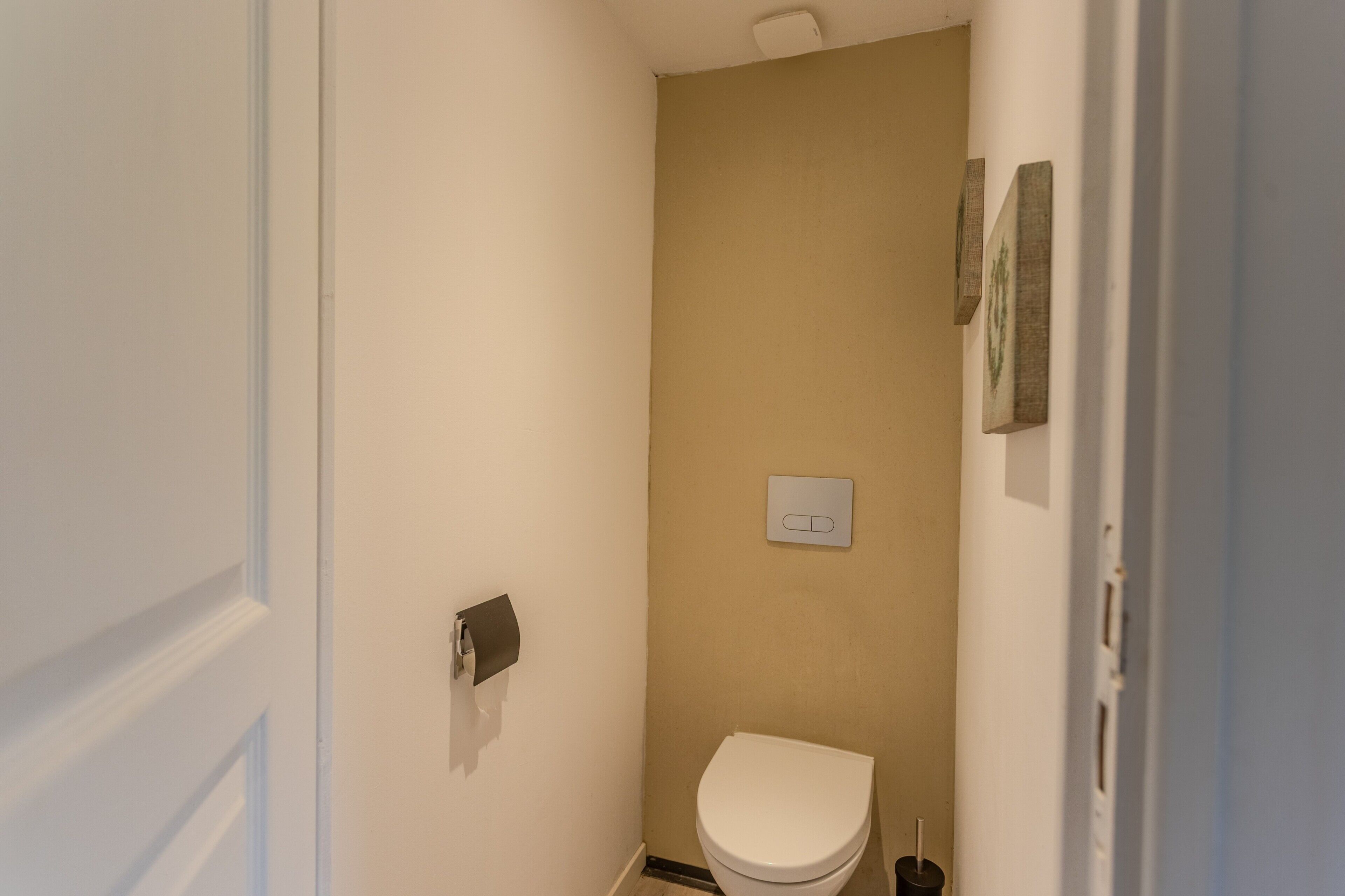 Een kleine, smalle badkamer met een toilet, een in de muur gemonteerde lichtschakelaar en een deur.