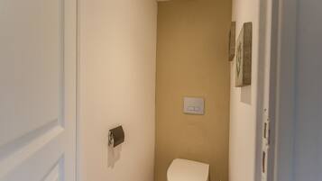 Een kleine, smalle badkamer met een toilet, een in de muur gemonteerde lichtschakelaar en een deur.