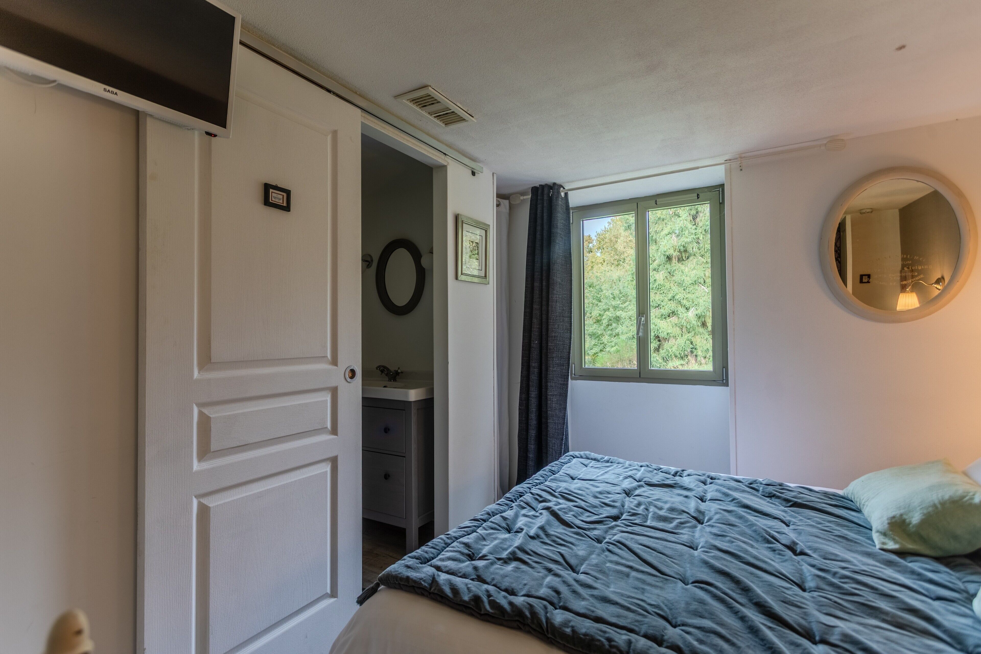 Een slaapkamer met een bed, een raam met uitzicht op groen, een ronde spiegel en een deur naar een badkamer.