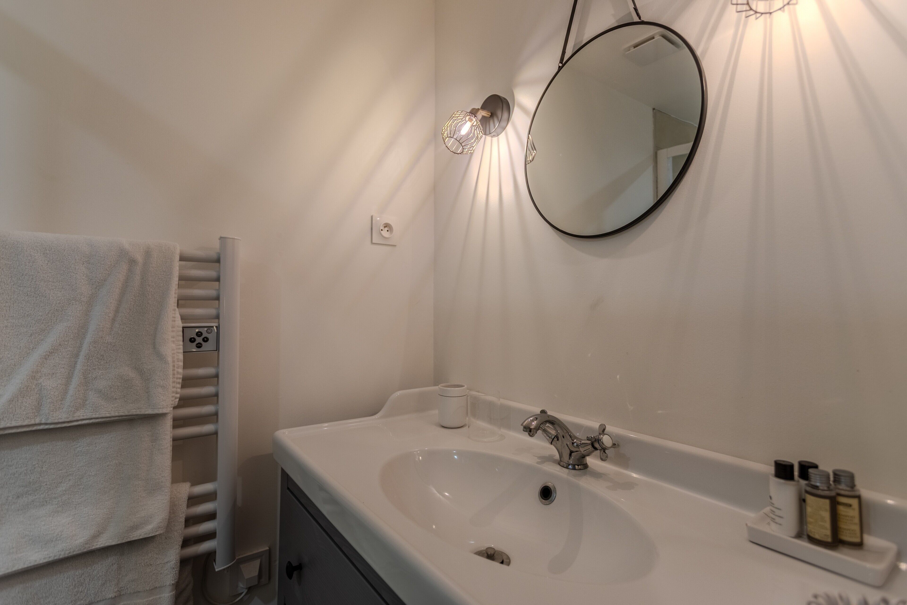 Ein Badezimmer mit einem weißen Waschbecken, einem runden Spiegel, einem Handtuchhalter und einer Leuchte.
