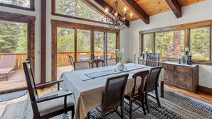 A dining area with a wooden table, chairs, and a chandelier. Large windows provide views of the surrounding forest. - Evergreen Hideout Retreat (Kings Beach)