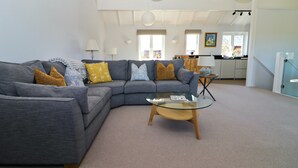A living room with a grey sectional sofa, a glass coffee table, and a kitchen in the background.