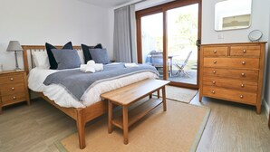 A bedroom with a bed, bedside tables, a wooden dresser, a bench, and a view of the outdoors through a sliding glass door.