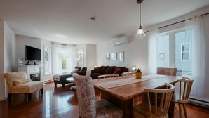 Een ruime woonkamer met een houten eettafel, zithoek, televisie en grote ramen.