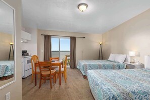 Et hotelværelse med to senge, et spiseområde med bord og stole, et tekøkken og et stort vindue med gardiner.