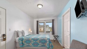 Ein Schlafzimmer mit Bett, Schreibtisch, Stuhl, einem an der Wand befestigten Fernseher und einem Fenster mit Vorhängen.