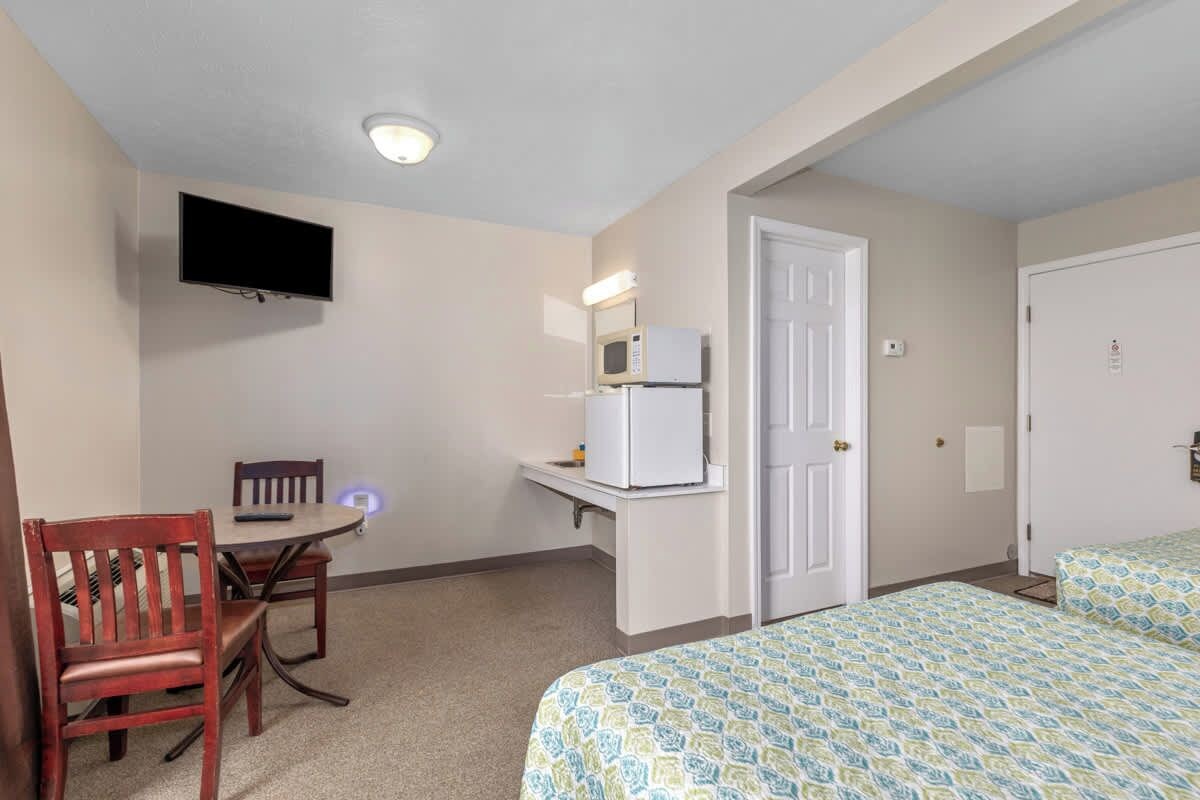 A hotel room with a bed, a desk with a microwave, a TV mounted on the wall, and a door.