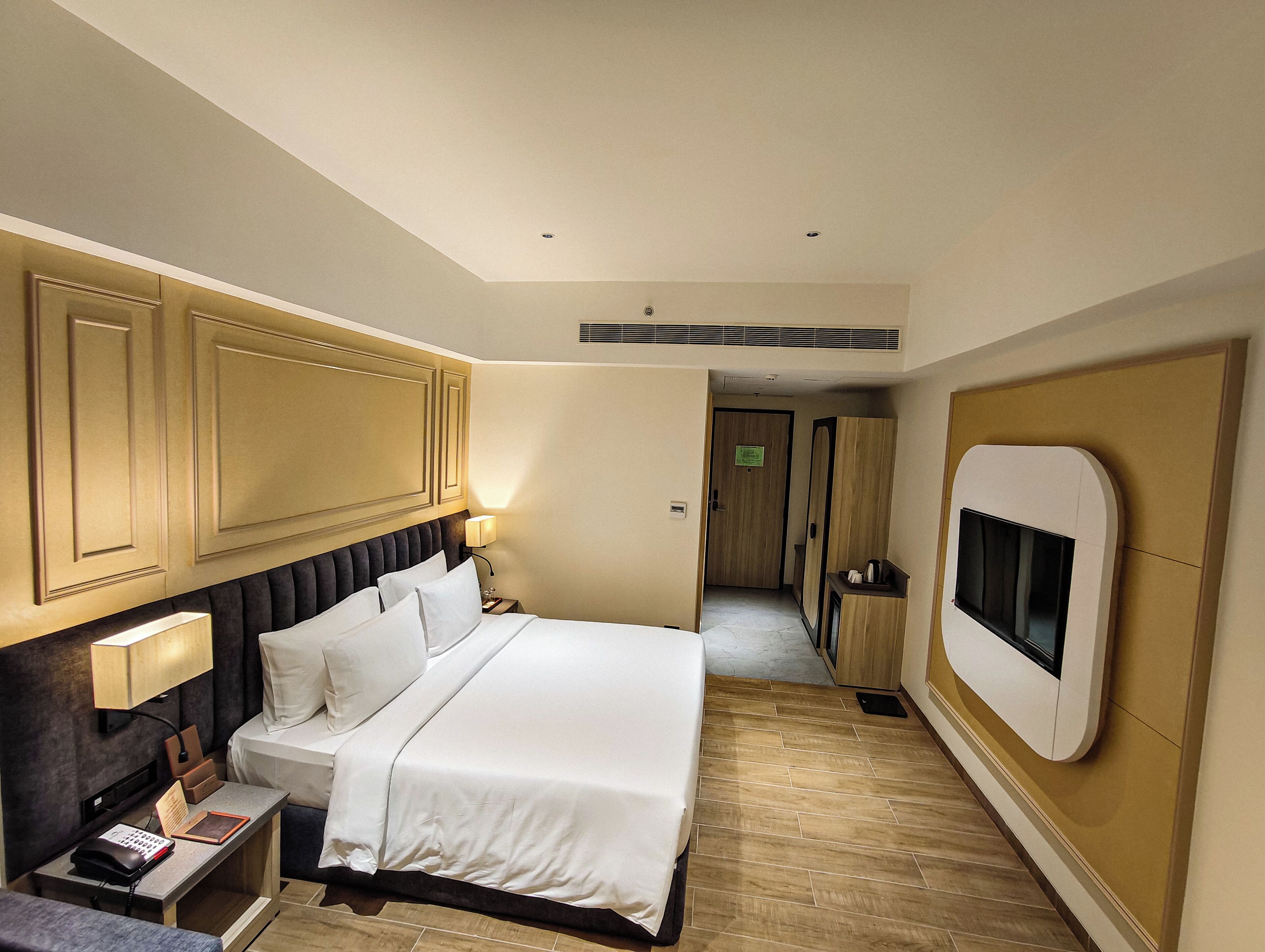 Un cuarto de hotel con una cama grande, una mesita de noche con lámpara, un televisor montado en la pared y piso de madera.