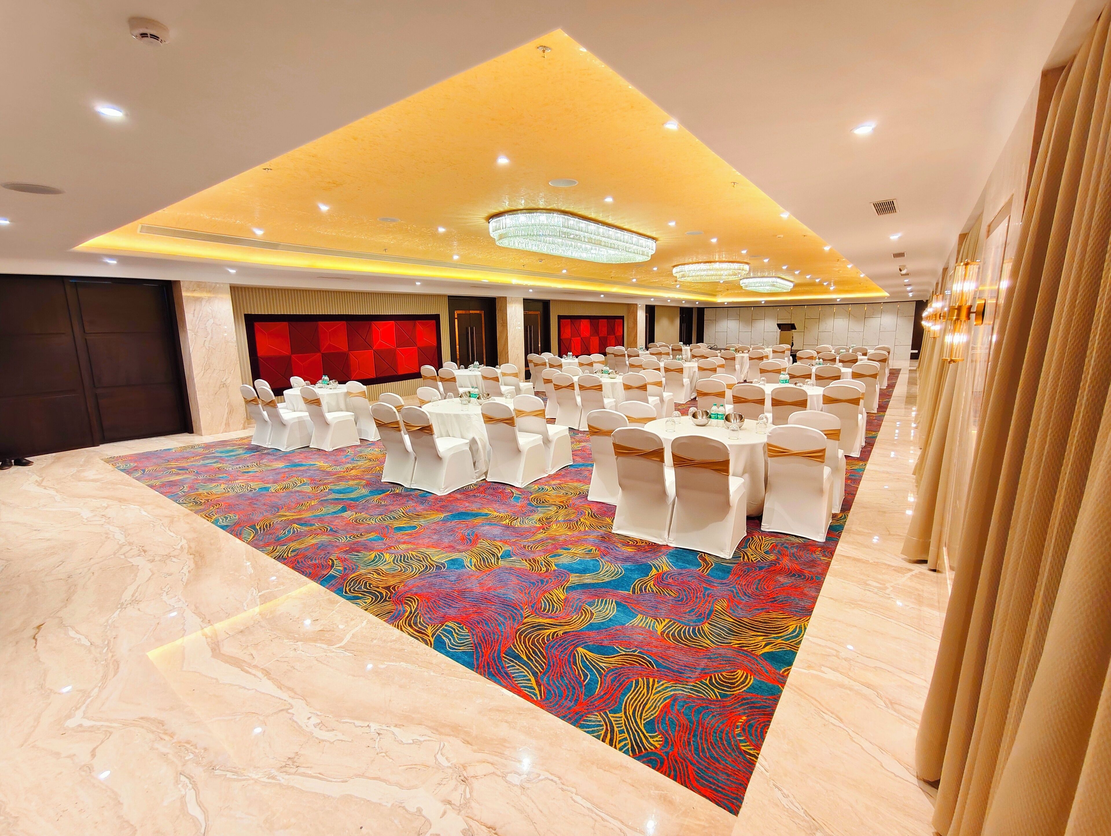 Une salle de banquet avec des tables rondes et des chaises blanches, une moquette colorée et un design de plafond moderne.