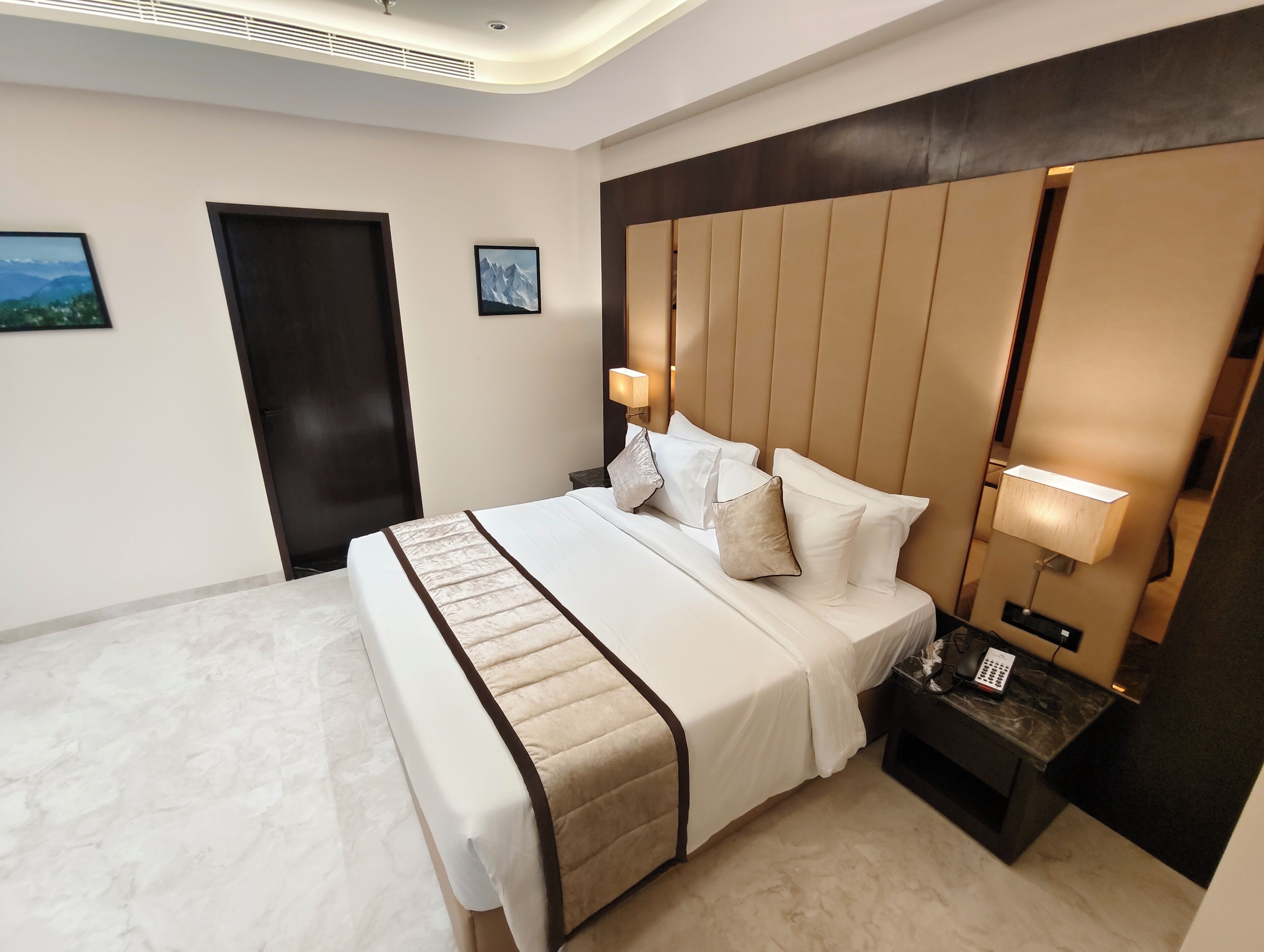 A hotel room with a large bed, a headboard with vertical panels, a bedside table with a lamp, and a door.