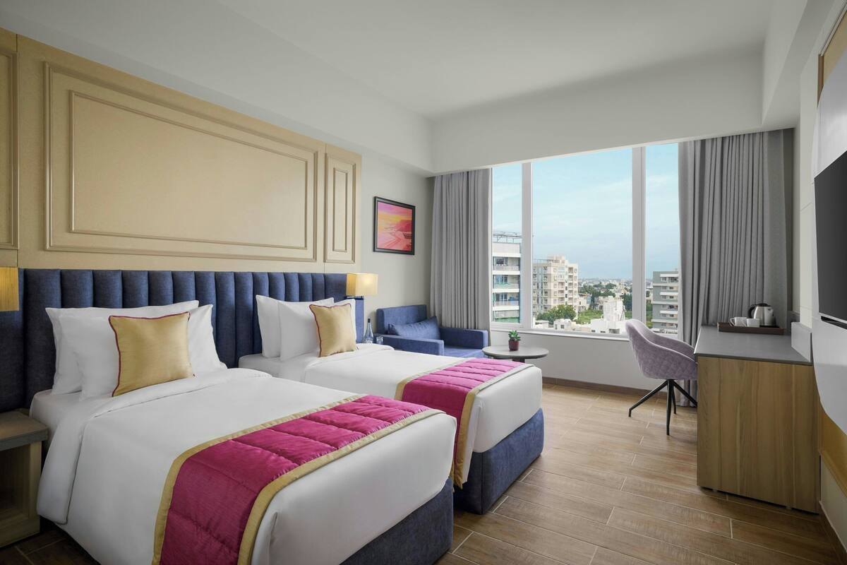 A hotel room with two beds, a desk, a chair, a TV, and a large window with a city view.