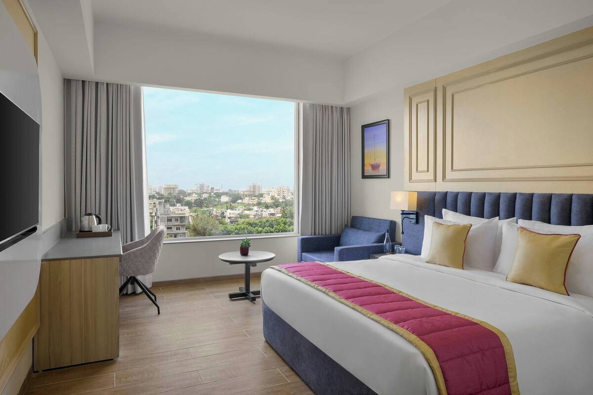 A hotel room with a large bed, a desk, a chair, a small table, and a view of the city.