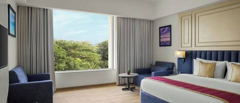 A hotel room with a large bed, a sofa, a small table, and a view of greenery outside.