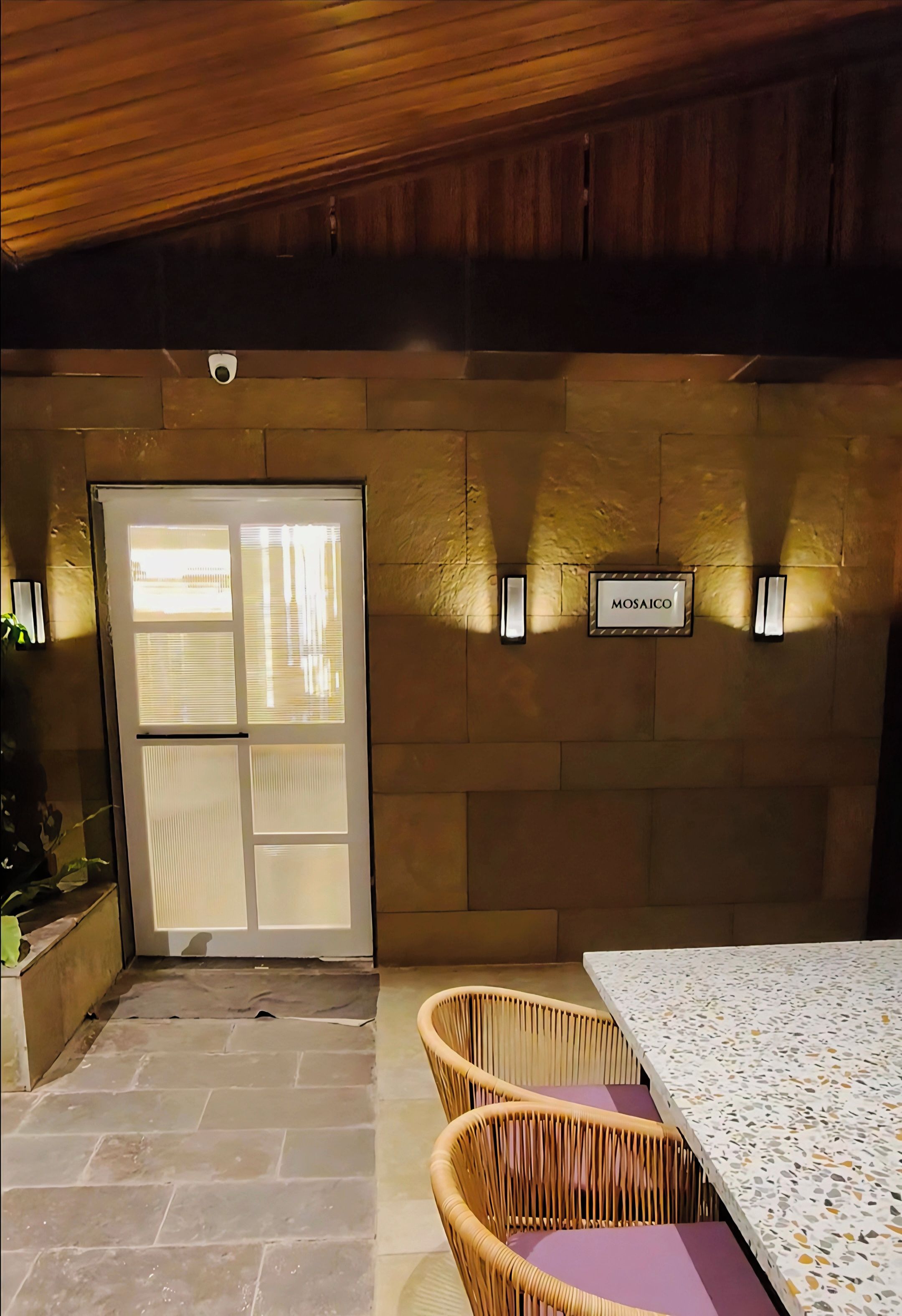 A room with a stone floor, a glass door, wicker chairs, a table with a patterned tablecloth, and a wall-mounted sign labeled "MOSAICO".