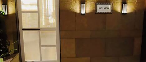 A room with a stone floor, a glass door, wicker chairs, a table with a patterned tablecloth, and a wall-mounted sign labeled "MOSAICO".