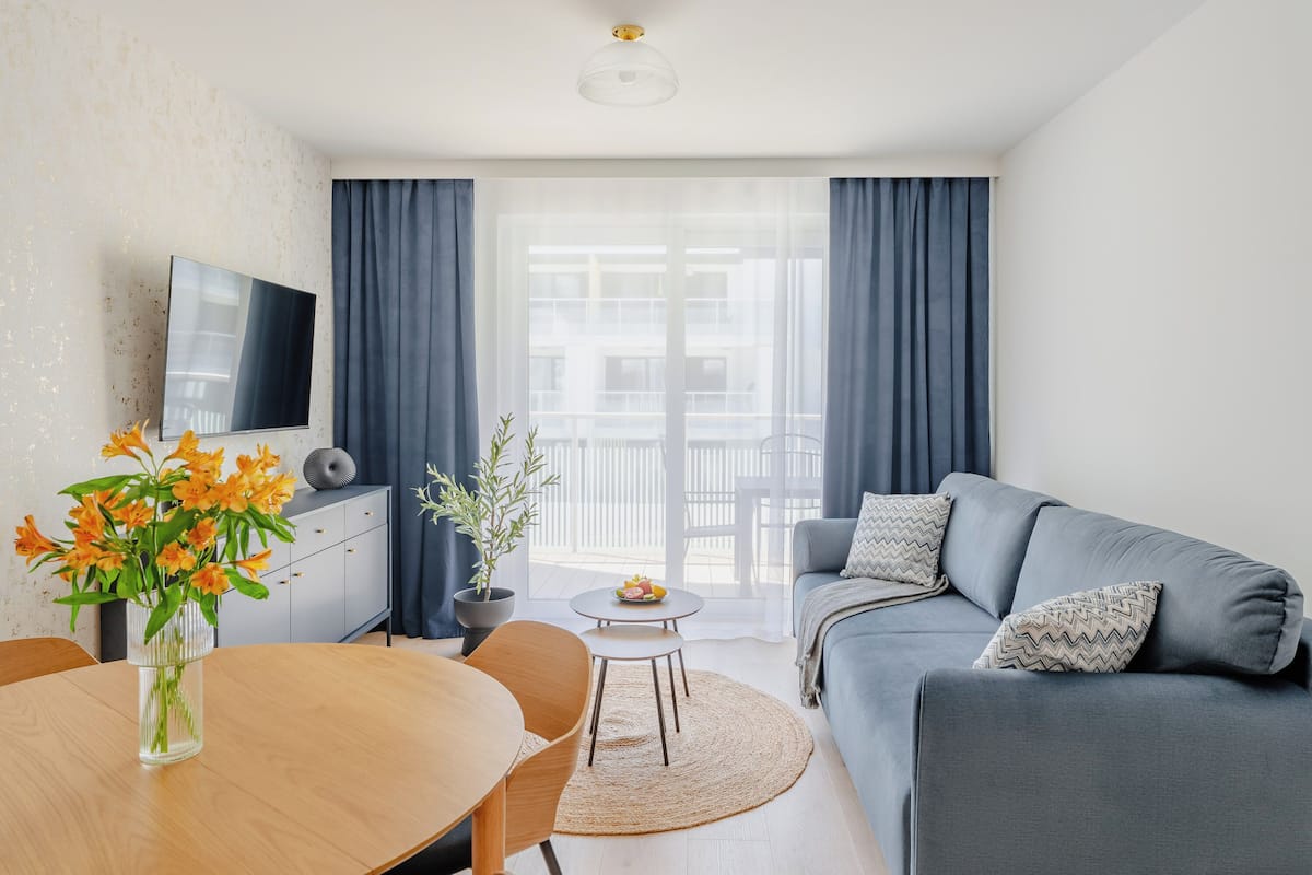 A modern living room with a sofa, a dining table with a vase of flowers, a small round table with a fruit bowl, and a TV mounted on the wall.