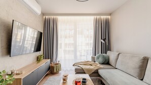 Egy modern nappali, melyben található egy síkképernyős televízió, egy kanapé, egy dohányasztal és egy étkezősarok, ahol egy tálca süteménnyel van.