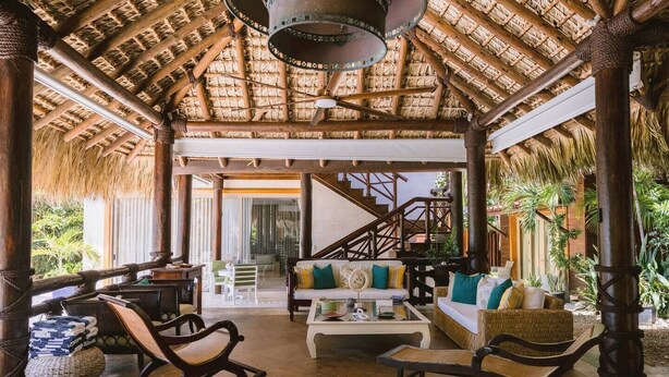 Un espacio interior amplio con techo de paja, muebles de madera y una escalera que lleva a un nivel superior.