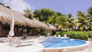 Un área de piscina con sillones de descanso, una estructura de techo de paja y palmeras.