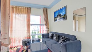 A living room with a sofa, ottoman, and coffee table, a large window with curtains, and a cityscape painting on the wall. - Hotel O Putra Family Homestay Sikunir (Kejajar)