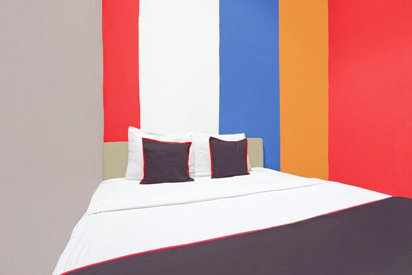 Una cama con ropa de cama blanca y dos almohadillas oscuras, frente a una pared con rayas verticales en rojo, blanco, azul y naranja.