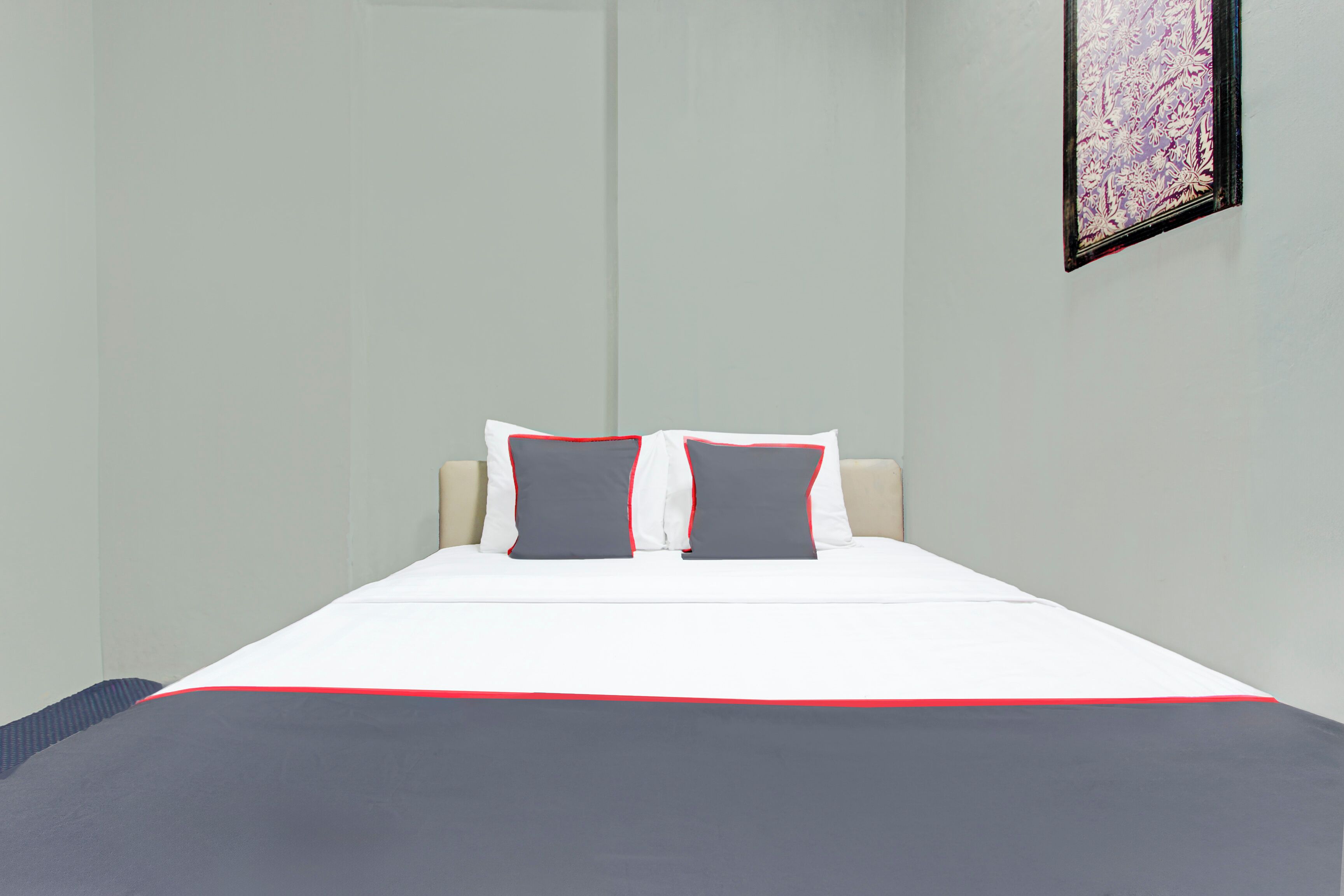 Cama bem arrumada com roupa de cama branca e cinza, dois travesseiros com detalhes vermelhos e um quadro na parede.