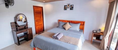 Un dormitorio con techo de madera, una cama con ropa de cama gris, una mesita de noche con lámpara, un escritorio de madera con espejo y un ventilador de pared.