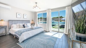 A modern bedroom with a large bed, a view of the outdoors, and a balcony with a potted plant. - Pickleball, Pool, Spa, Casita, Firepit, Gameroom (Desert Hot Springs)