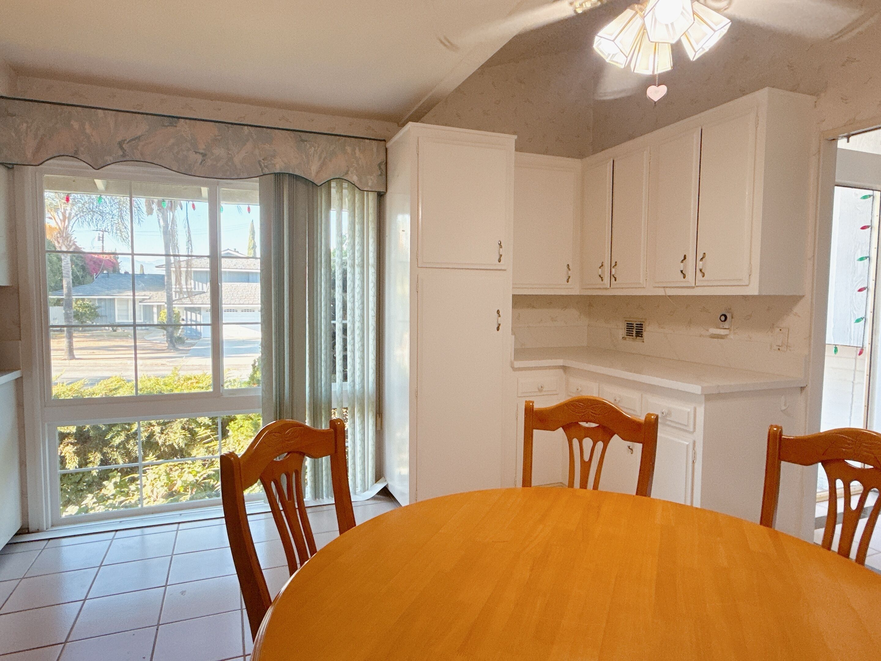 Comedor con una mesa redonda y cuatro sillas de madera, armarios blancos y vistas al exterior a través de una ventana con cortinas.