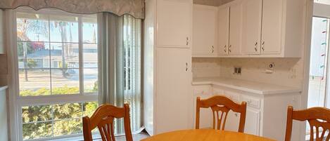 Comedor con una mesa redonda y cuatro sillas de madera, armarios blancos y vistas al exterior a través de una ventana con cortinas.