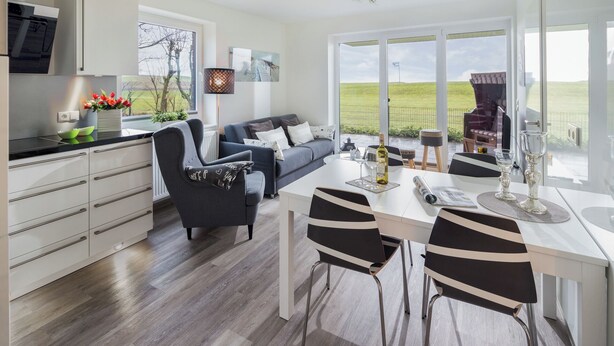 A modern kitchen with white cabinets, a dining area with a white table and black chairs, and a large window offering a view of a green field. - // HOUSE DEICHBLICK - APARTMENT 1 // (Norderney)