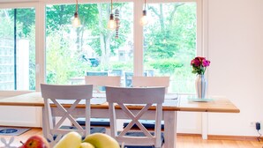 A dining area with a wooden table and chairs, a vase with flowers, and a view of greenery outside.