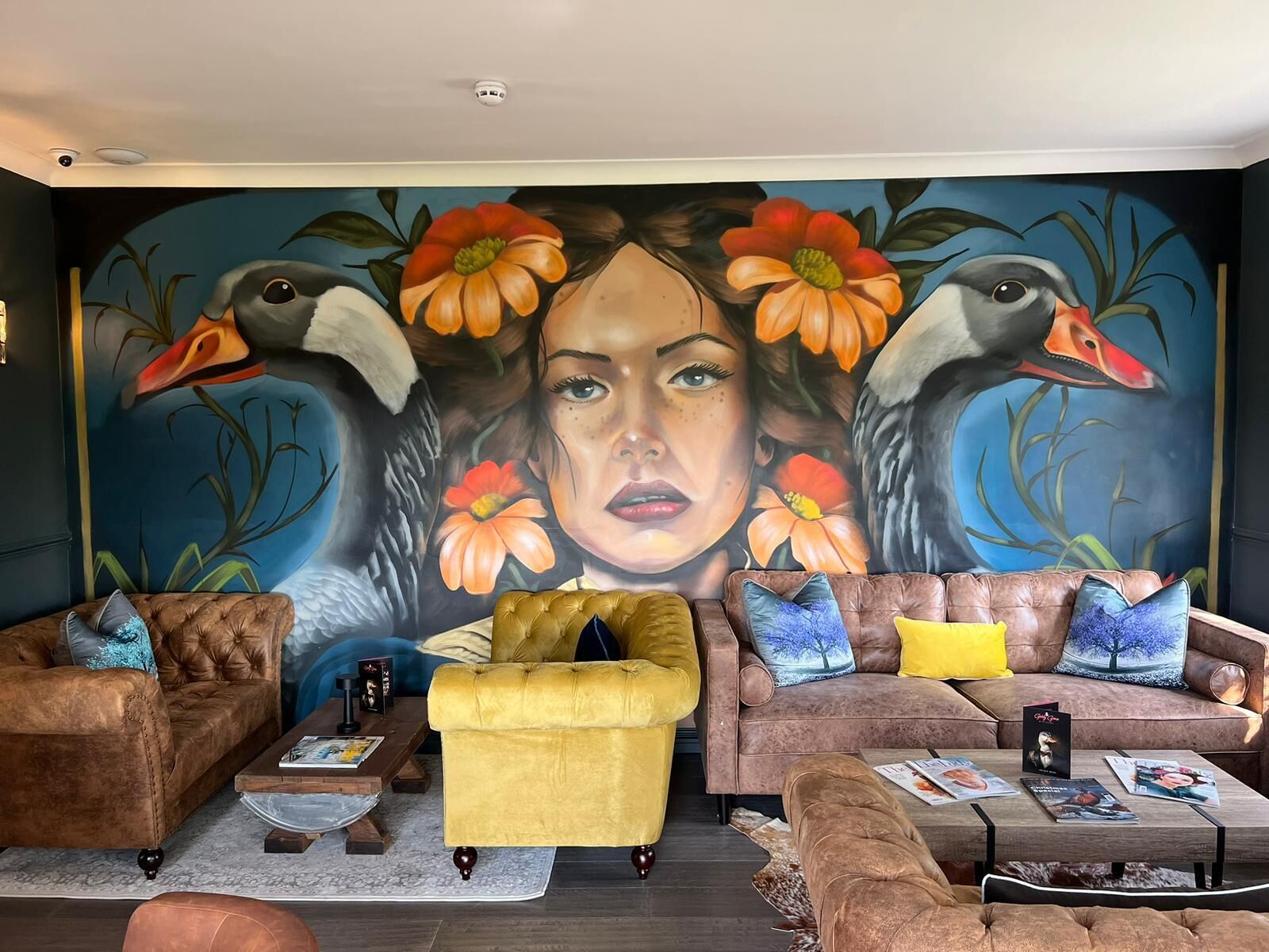 Un salon agrémenté d’une grande fresque murale, de deux canapés, d’un fauteuil jaune et d’une table basse sur laquelle sont posés des magazines.