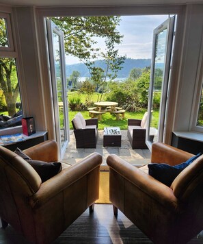 A room with a view of a garden and mountains through large windows. There are two leather chairs and a small table in the center. - The Ryebeck Hotel (Bowness-on-Windermere)