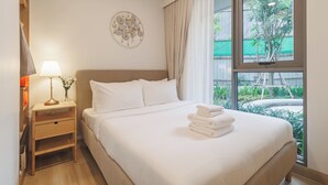 A hotel room with a bed, a nightstand with a lamp, a wall decoration, and a view of greenery outside. - Laguna SkyPark new brand apartments (Choeng Thale)