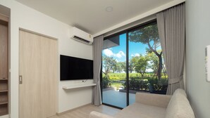 A modern living room with a flat-screen TV mounted on a white wall, a beige sofa, and a sliding glass door leading to a balcony with a view of greenery. - Laguna SkyPark new brand apartments (Choeng Thale)