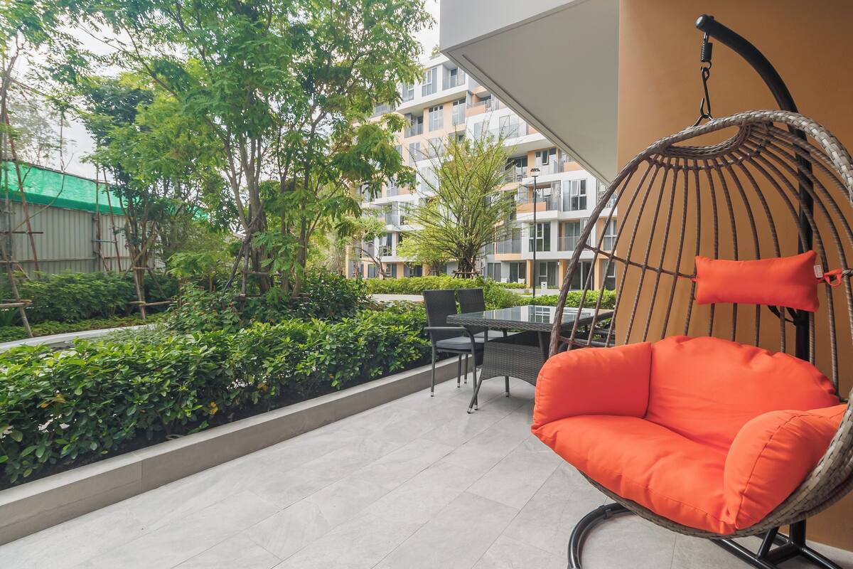 Una silla colgante con cojines anaranjados, una mesa con sillas y vistas a un edificio con balcones y vegetación.