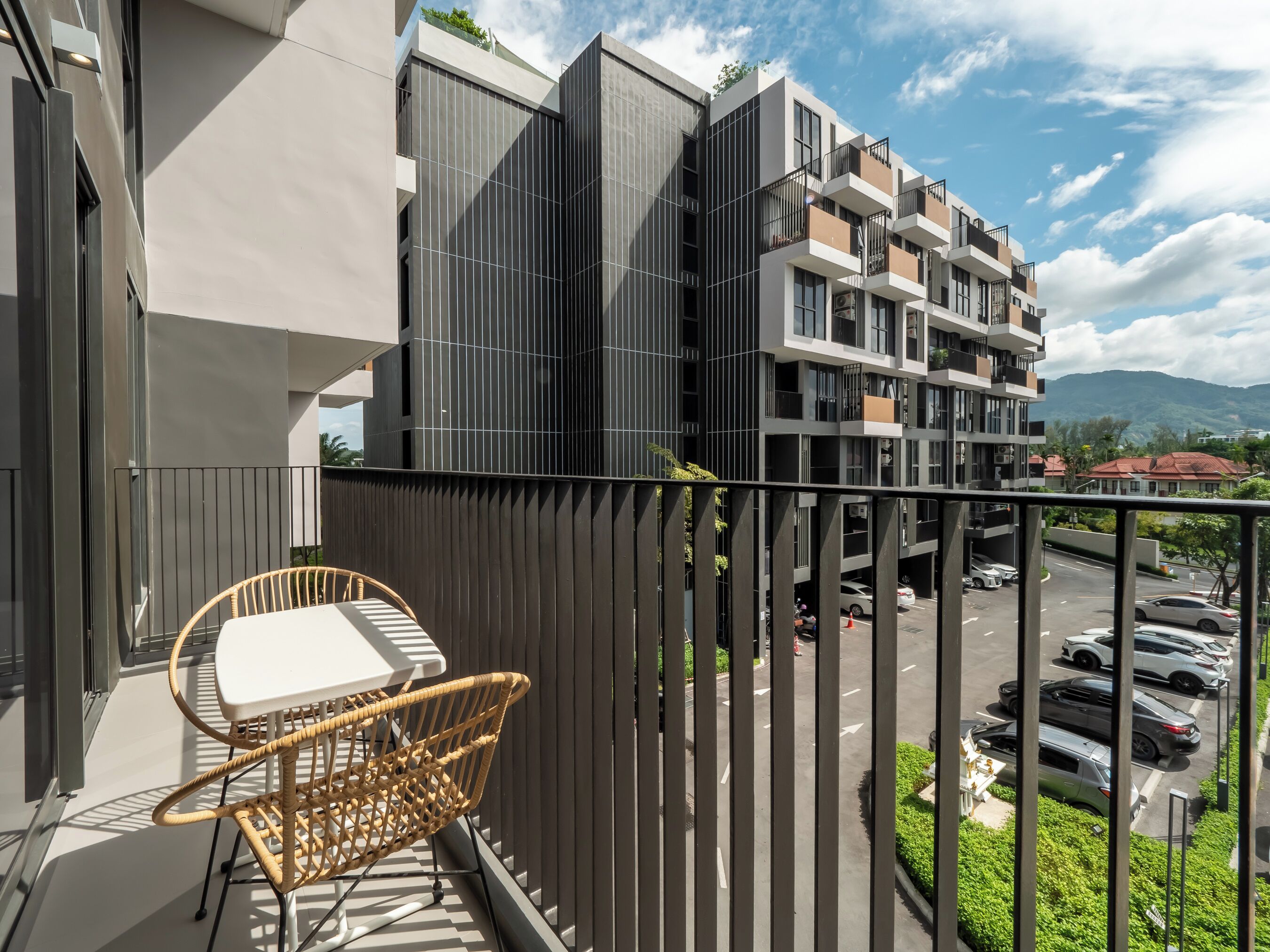 A balcony with a wicker chair and table, overlooking a parking area and modern buildings.