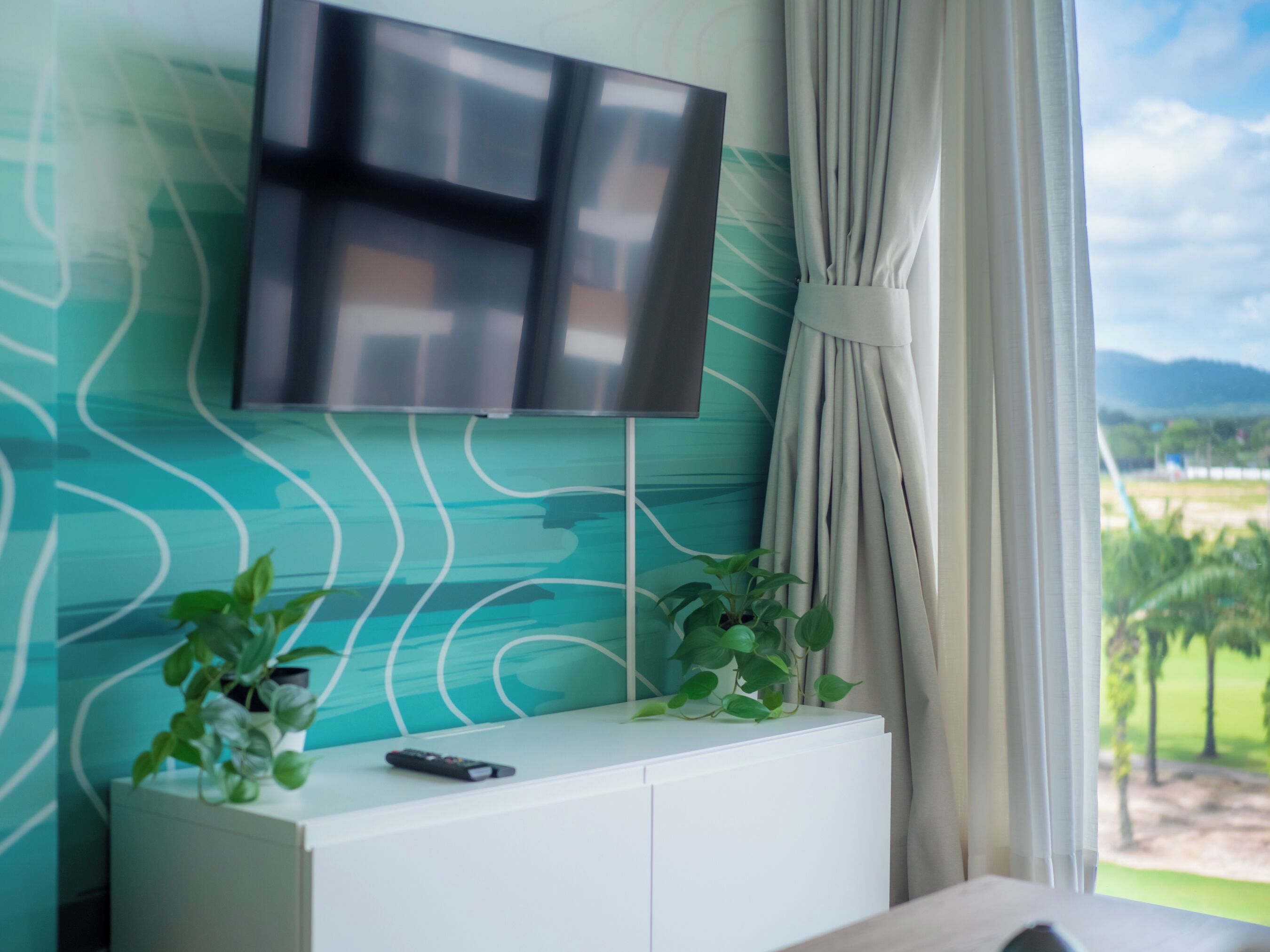 Une pièce dotée d’un téléviseur à écran plat fixé au mur, lequel présente un motif ondulé, d’un meuble blanc avec une télécommande, et d’une fenêtre avec des rideaux.