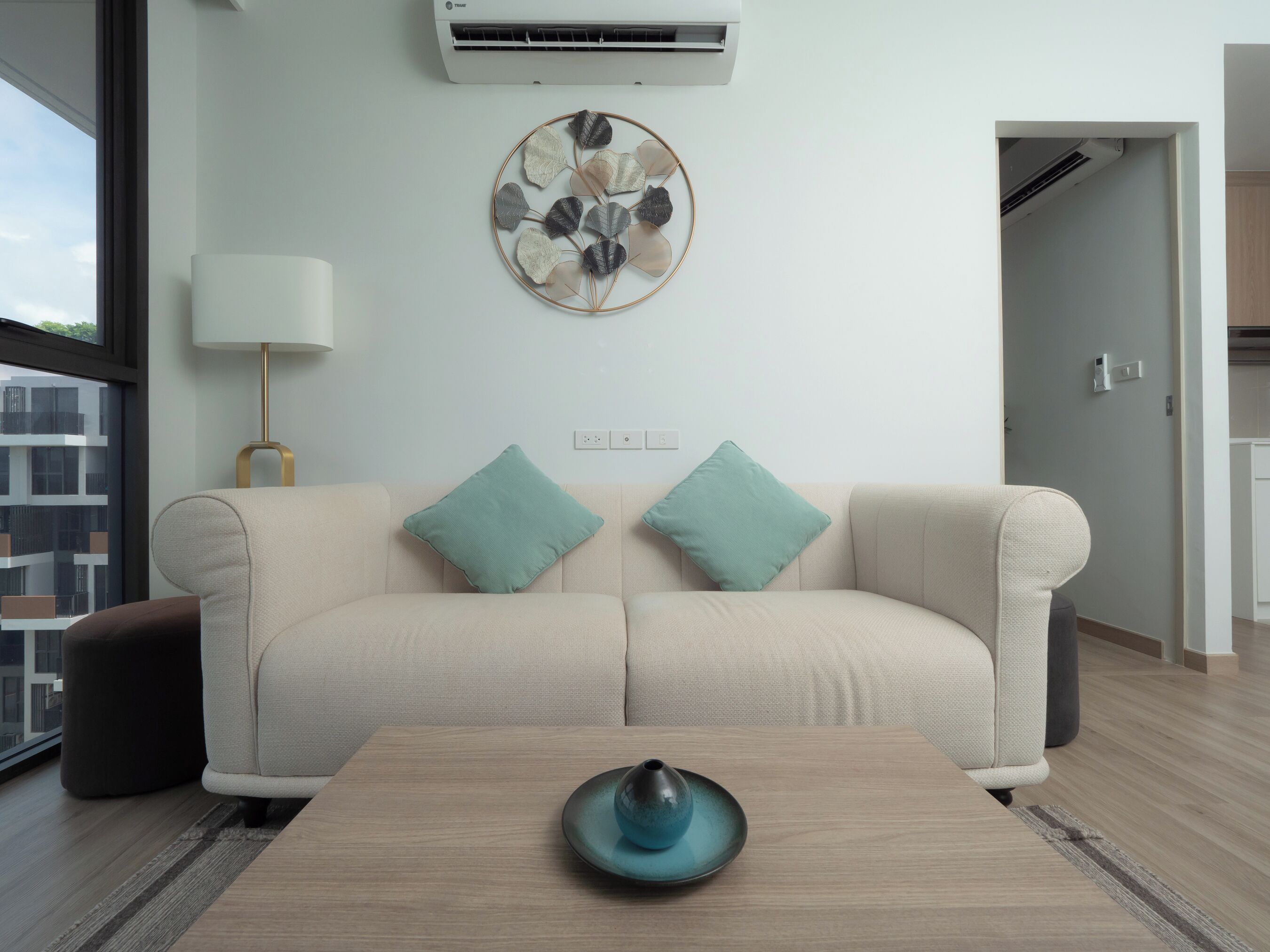 Un salón moderno con un sofá color beige, cojines turquesa, una mesa de centro de madera con un jarrón azul, un aire acondicionado instalado en la pared y una obra de arte con hojas decorativas.